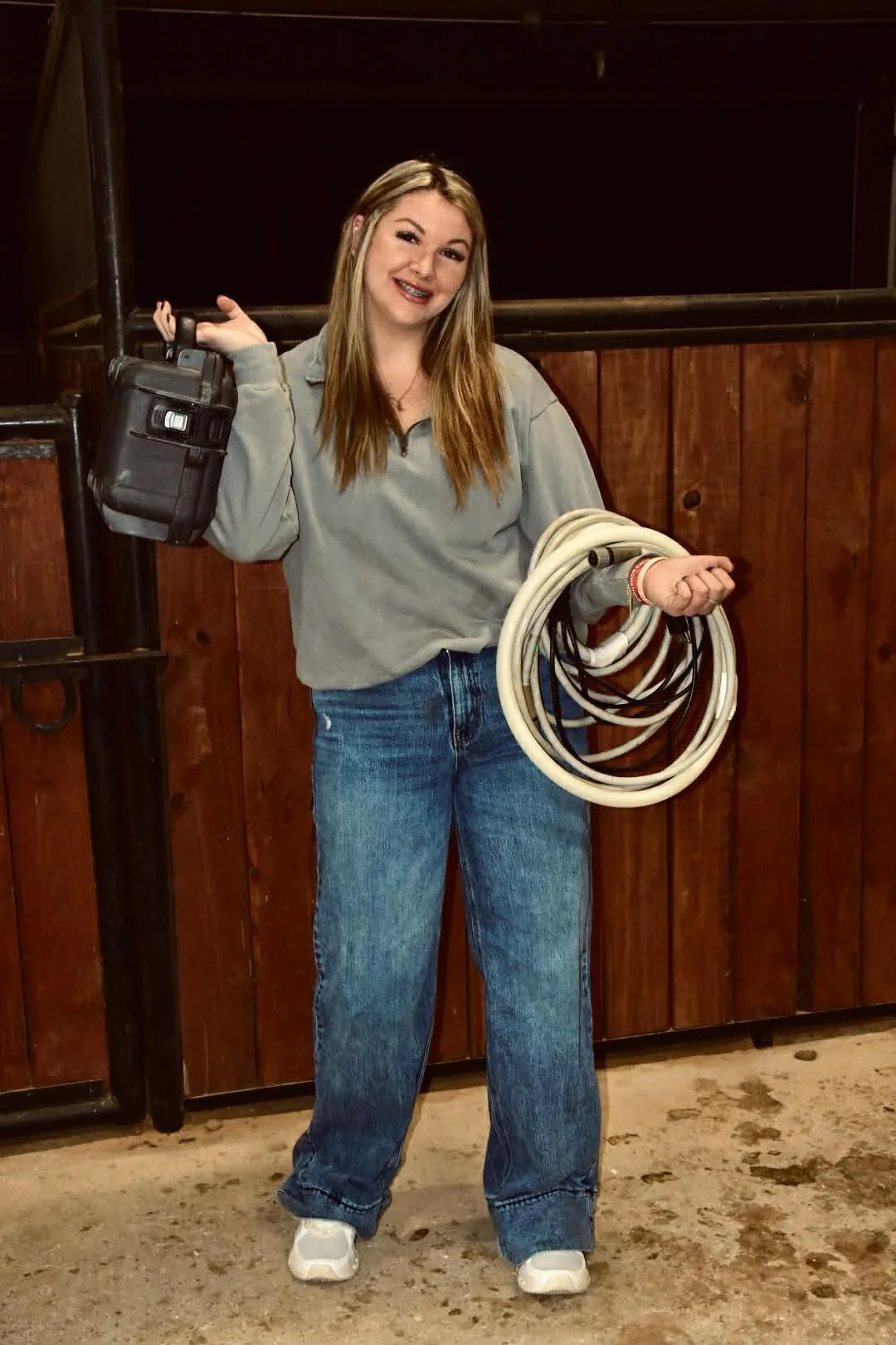 A young woman with long light brown hair, wearing a gray sweatshirt and blue jeans, standing in front of wooden barn walls, holding a coiled white cable in her left hand and a black toolbox in her right hand, smiling at the camera.