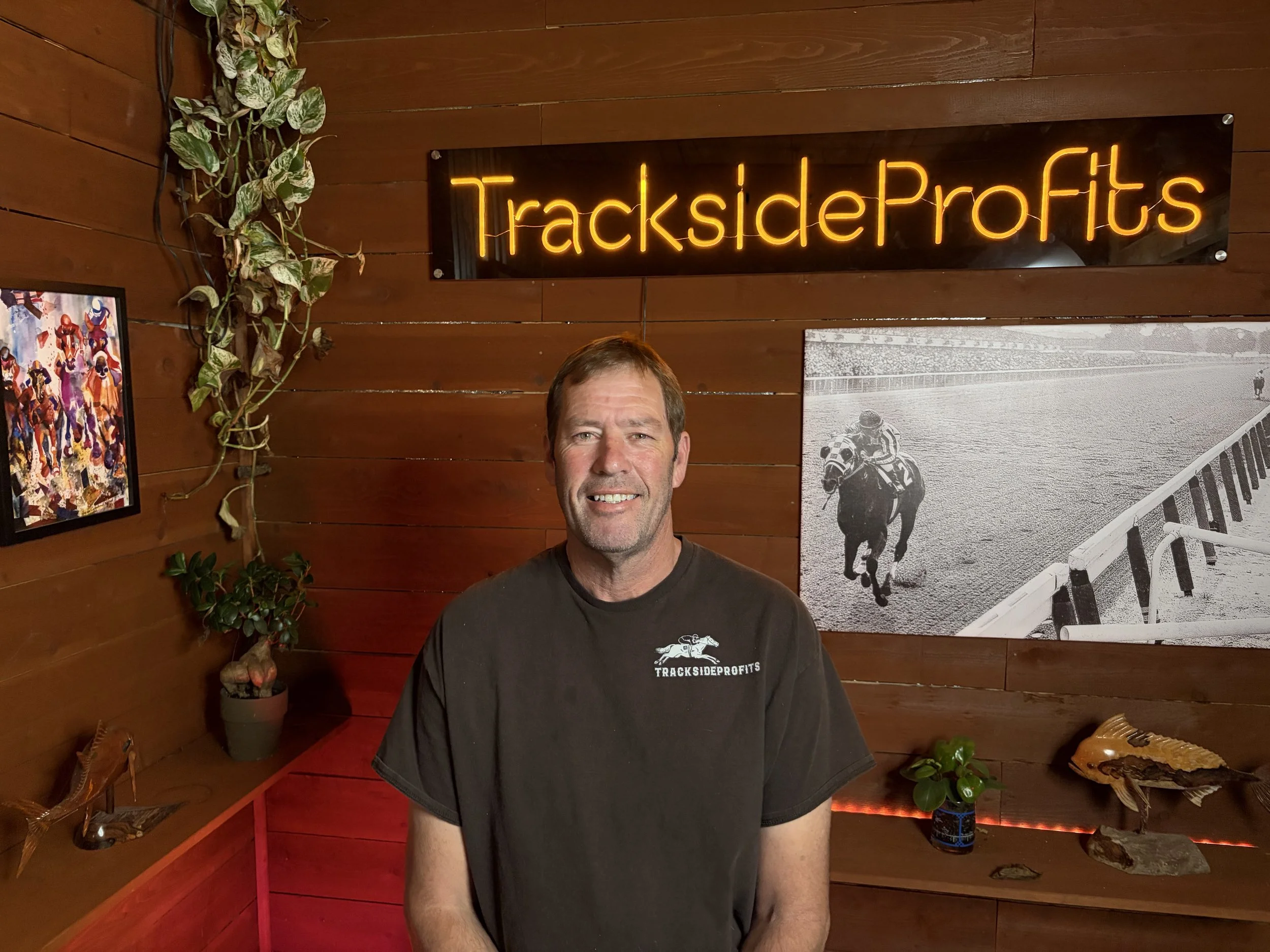 A smiling man in a black TracksideProfits t-shirt stands in a wooden interior next to plants and artwork, with a neon sign reading 'TracksideProfits' on the wall and a black-and-white horse racing photo in the background.