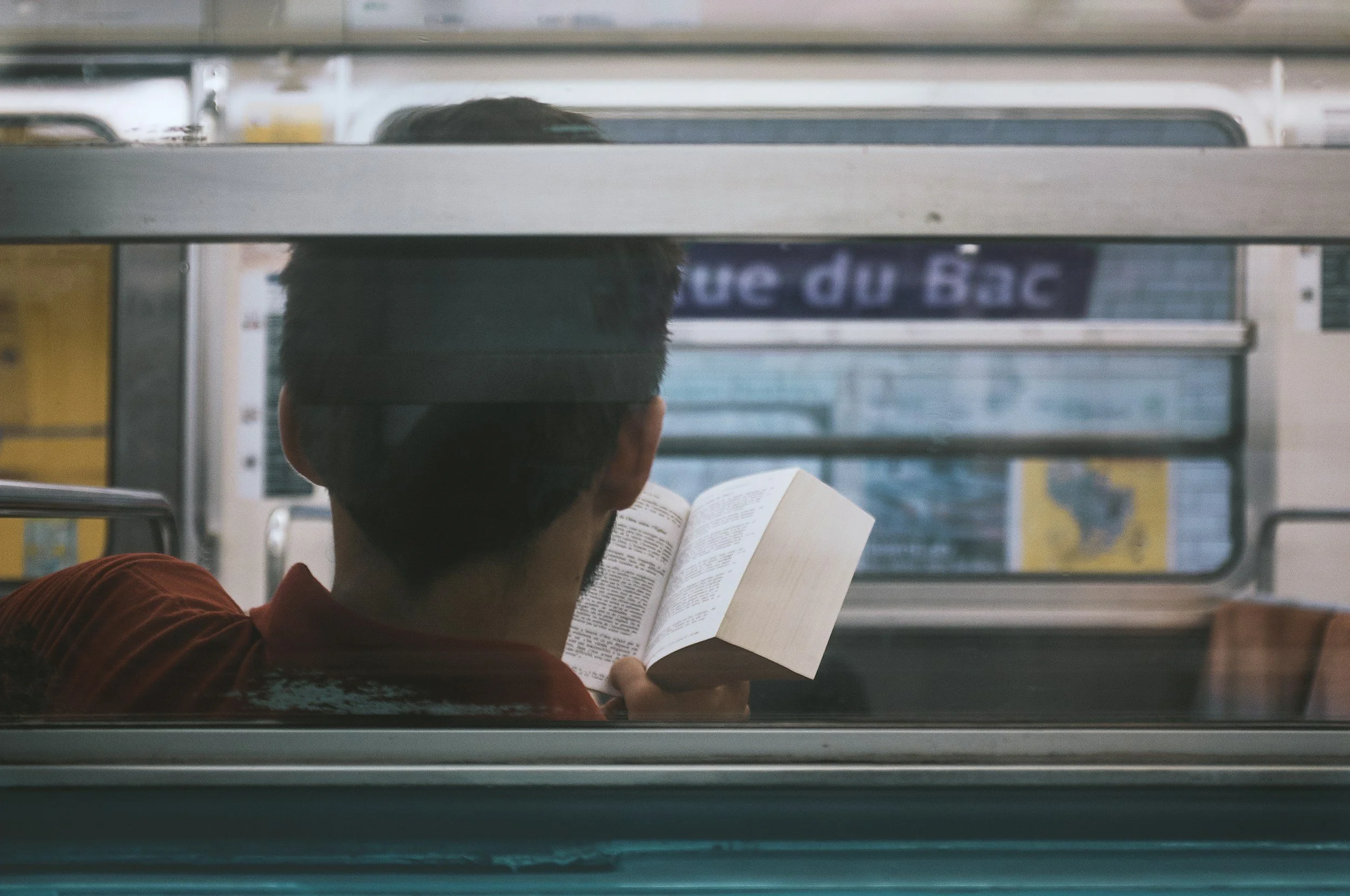 A person sitting on a bus, reading a book, with their back facing the viewer, through the window.