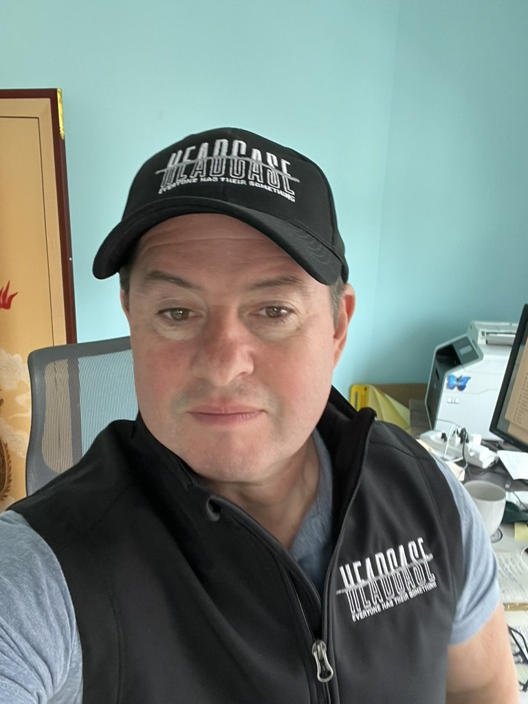 Chris K. Jones taking a selfie in an office. He is wearing a black cap and a matching vest both with the 'HEADCASE' logo.