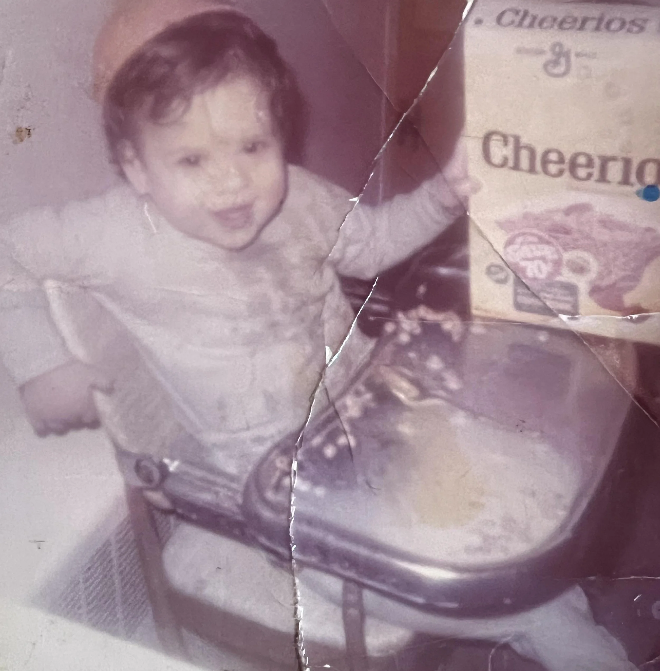 A young child with curly hair sitting at a table, looking up at the camera, with a box of Cheerios cereal nearby.