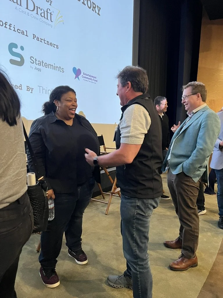 People standing in a conference room engaging in conversation, smiling, with a large screen in the background displaying logos and text.