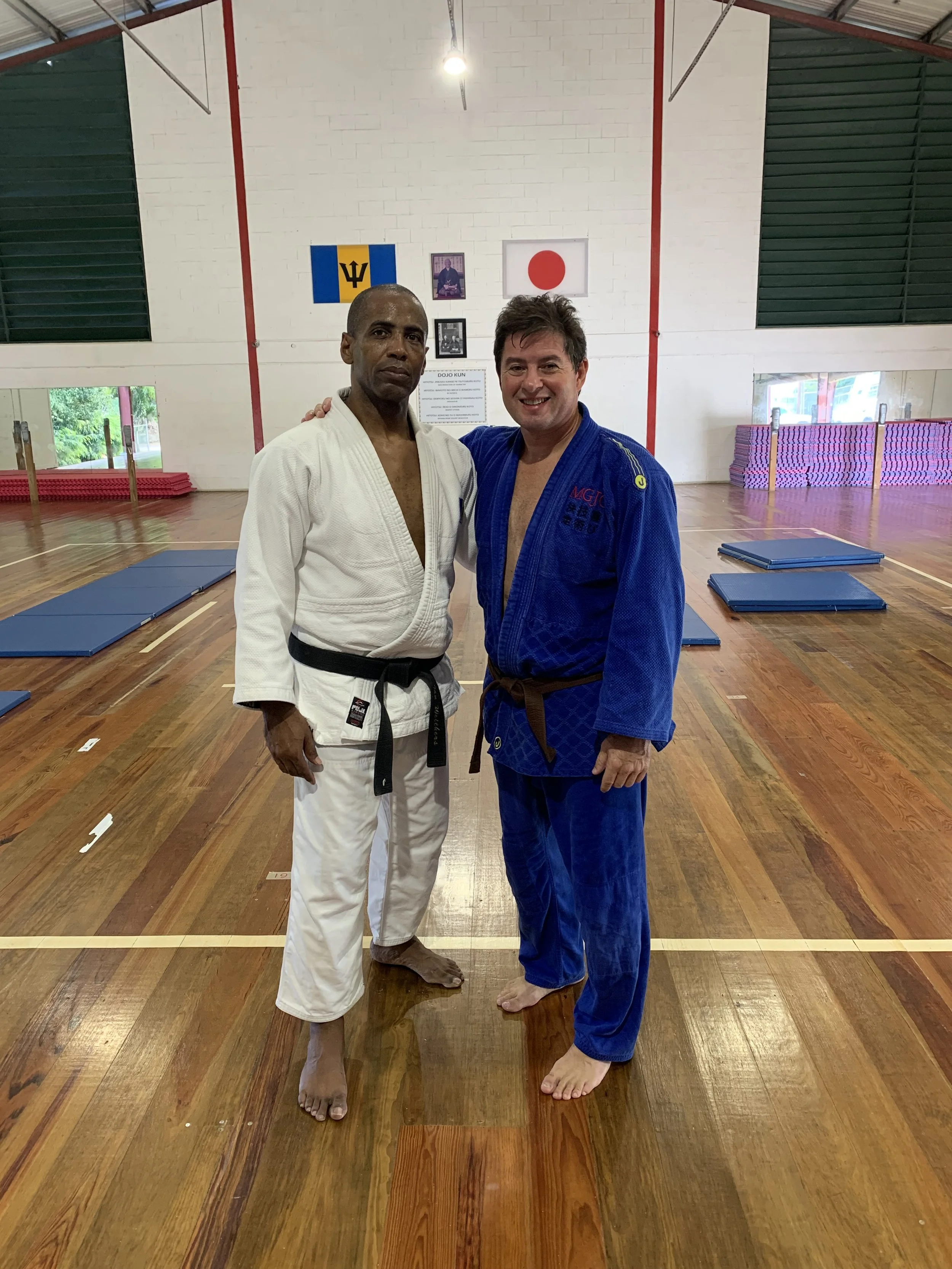 Two men in judo uniforms, one in white and the other in blue, standing together on a wooden floor inside a dojo. They are smiling and posing for the photo with their arms around each other's shoulders.