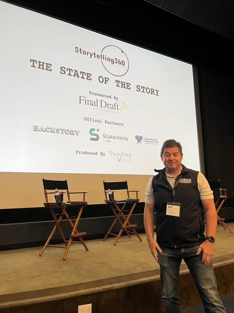 The writer, Chris K. Jones, standing in front of a presentation screen at a conference, with three empty chairs and water bottles on the stage. The screen displays the title 'The State of the Story' and logos of various sponsors.