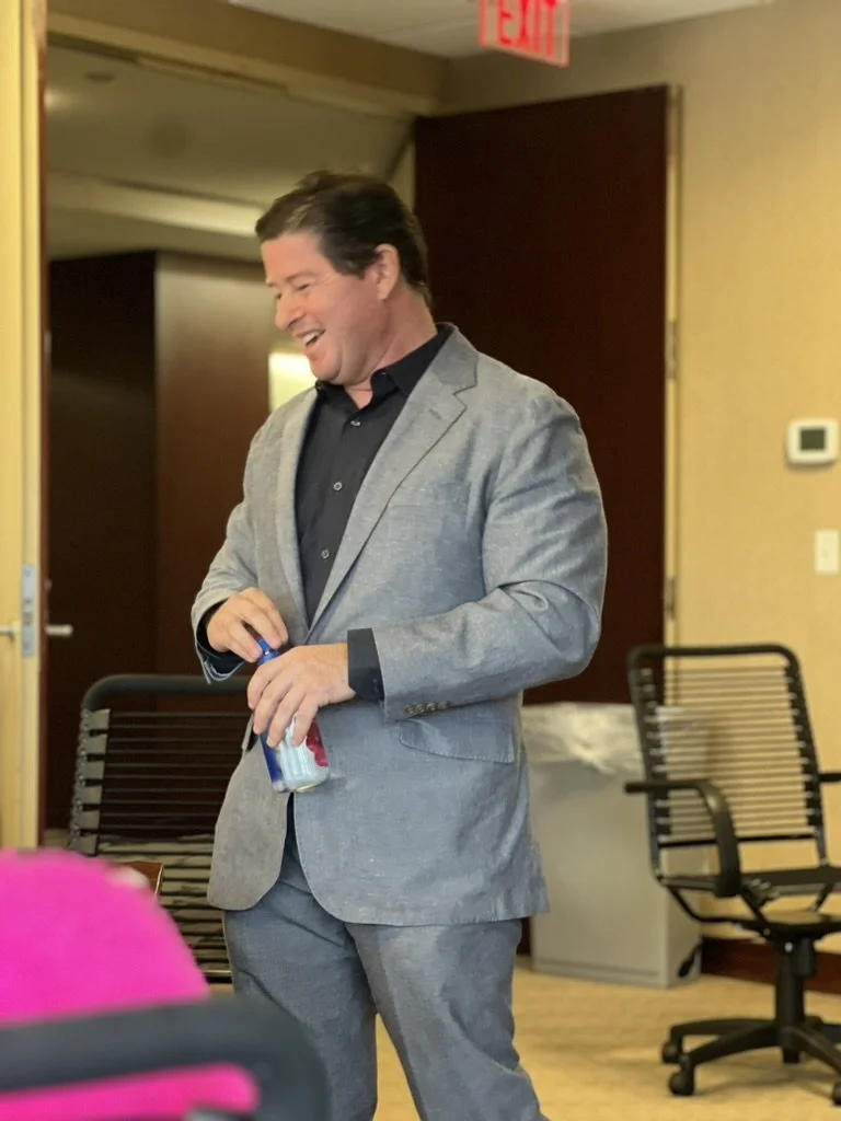 Chris K. Jones in a gray suit smiling and holding a canned beverage in a room with beige walls, black chairs, and an exit sign overhead.