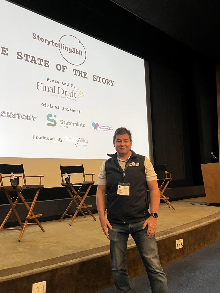 Chris K. Jones standing on a stage in front of a large screen during a conference or seminar. The screen displays logos and text related to storytelling and media companies. The man is smiling, wearing a vest with a badge labeled "1st".