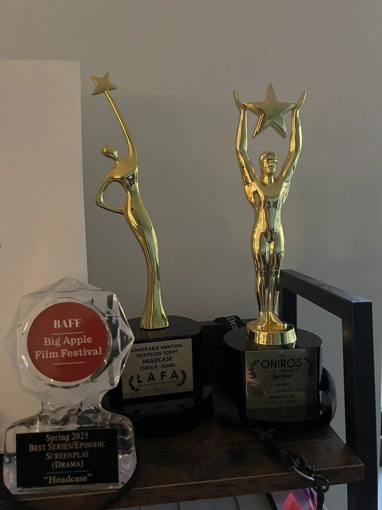 Three awards displayed on a dark wooden surface, including a glass trophy from the Big Apple Film Festival and two golden trophies for headscapes, one from LAFA and one from ONIROS.