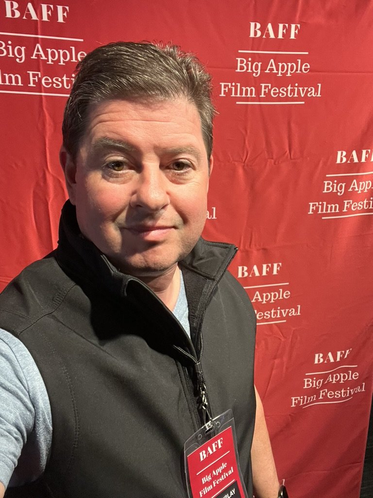 Chris K. Jones taking a selfie at the Big Apple Film Festival, standing in front of a red backdrop with the festival's name printed repeatedly.