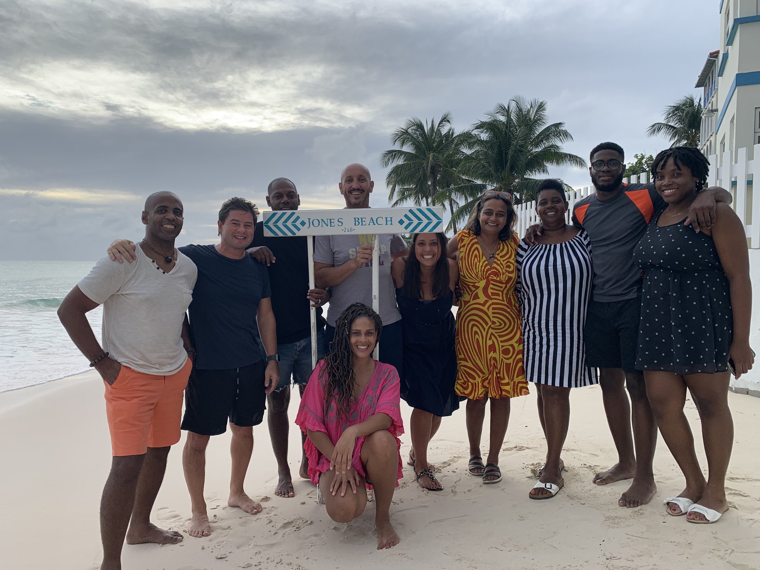 A diverse group of people standing on a beach under a cloudy sky, smiling and posing for a photo, with a sign that says 'Jones Beach' and palm trees in the background.