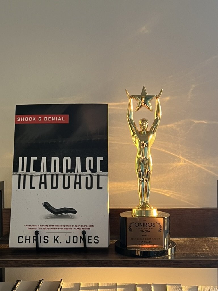 A trophy with a figure holding a star on top, placed beside a book titled 'Headcase' by Chris K. Jones, on a shelf with a plain background.