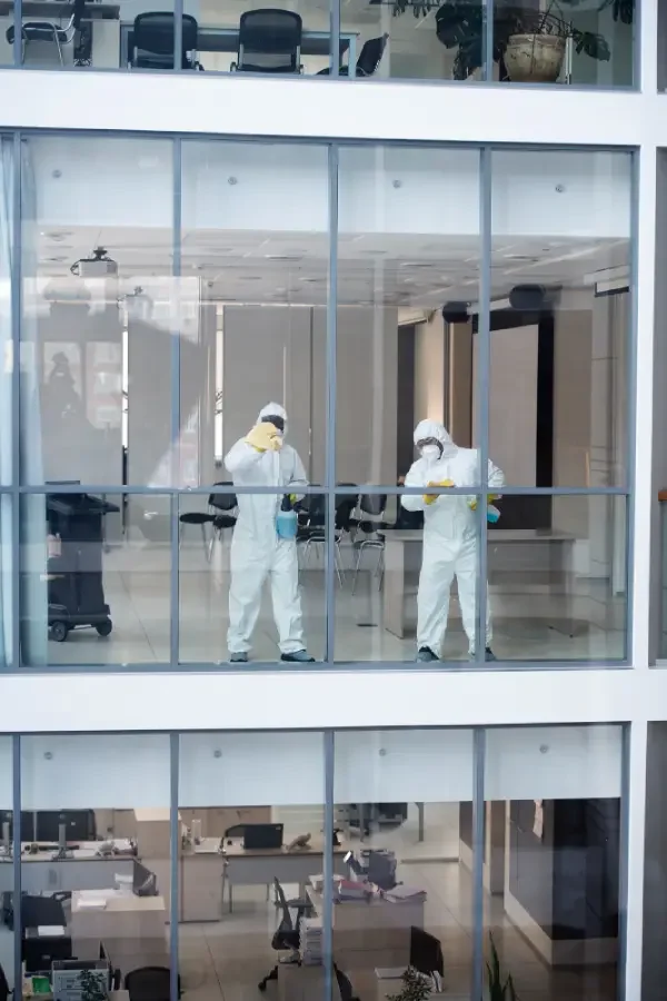 Two people wearing white hazmat suits, face masks, and gloves inside a glass-walled office, working with lab equipment.