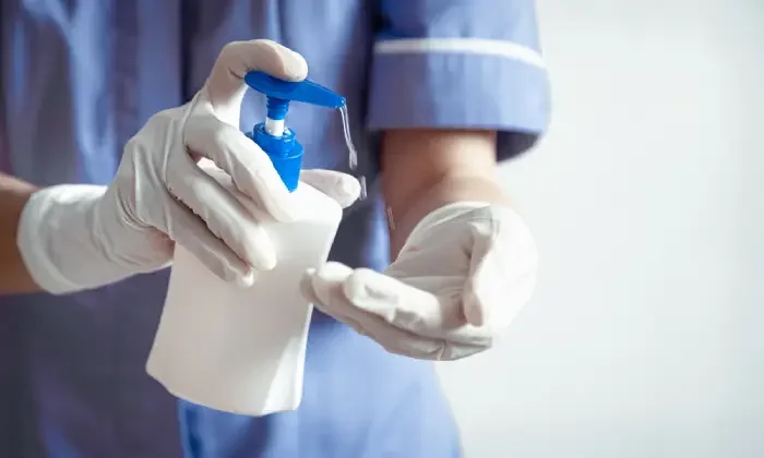 Person wearing gloves and blue medical scrubs pressing hand sanitizer dispenser.