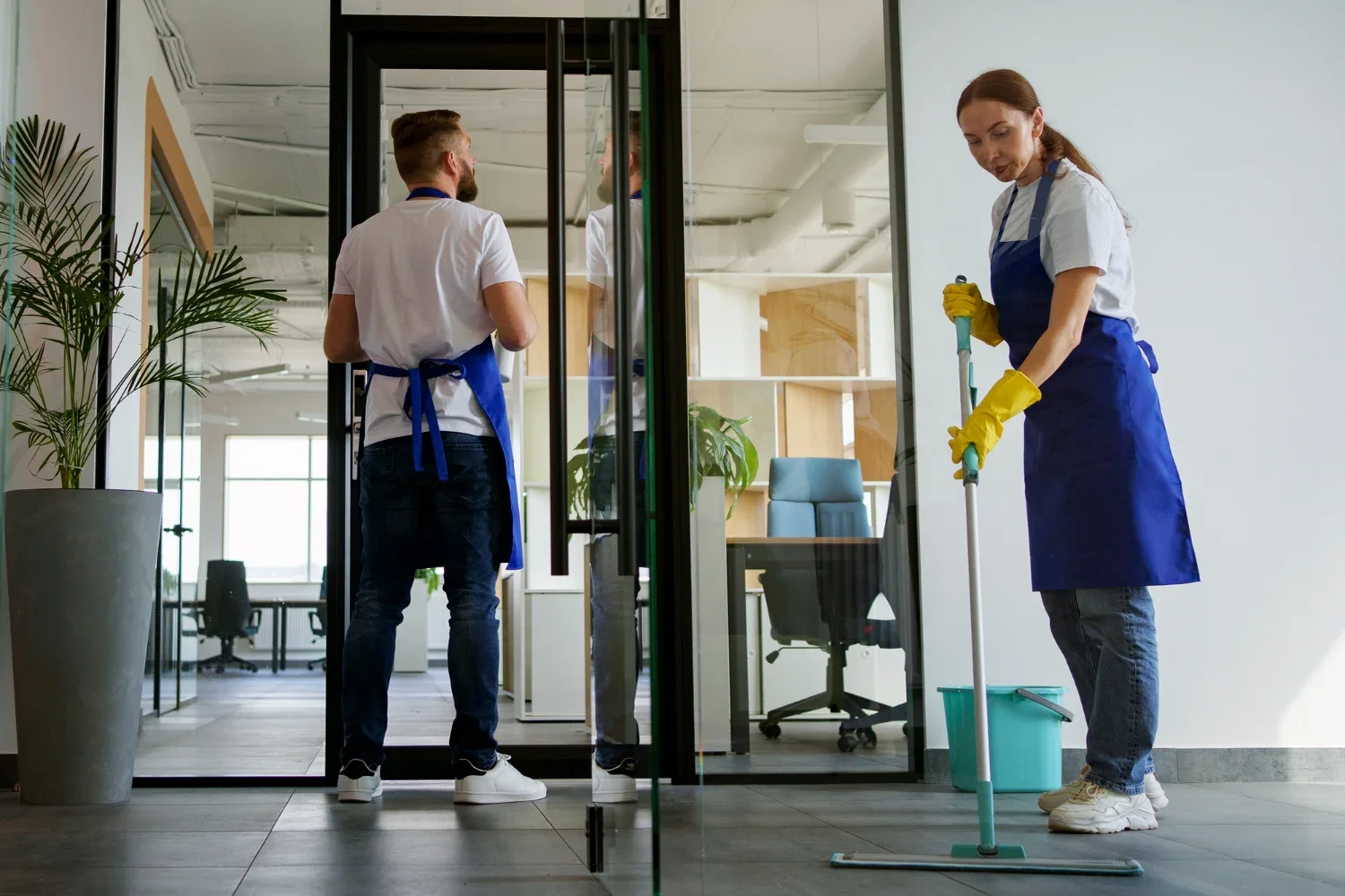 Two cleaning workers, a man and a woman, cleaning the glass doors of an office. The woman is mopping the floor, wearing yellow gloves and a blue apron. The man is facing away, wearing an apron and holding cleaning supplies, looking into the office.