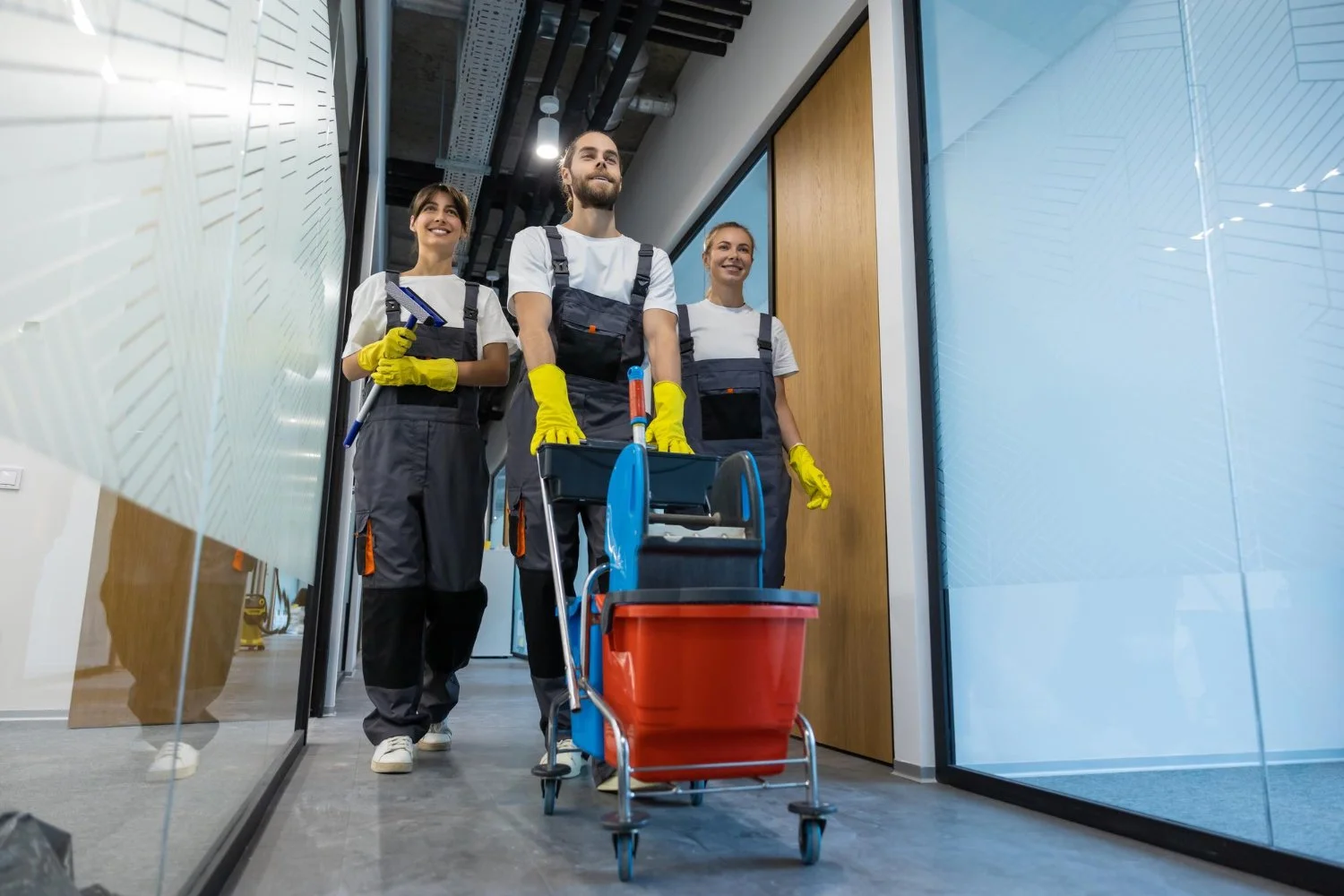 Three restaurant cleaning staff wearing gray uniforms and yellow gloves pushing a cleaning cart down a hallway.