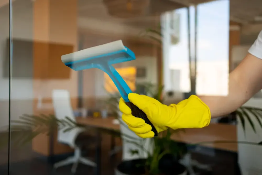 Person wearing yellow gloves cleaning glass window with squeegee in an office setting.