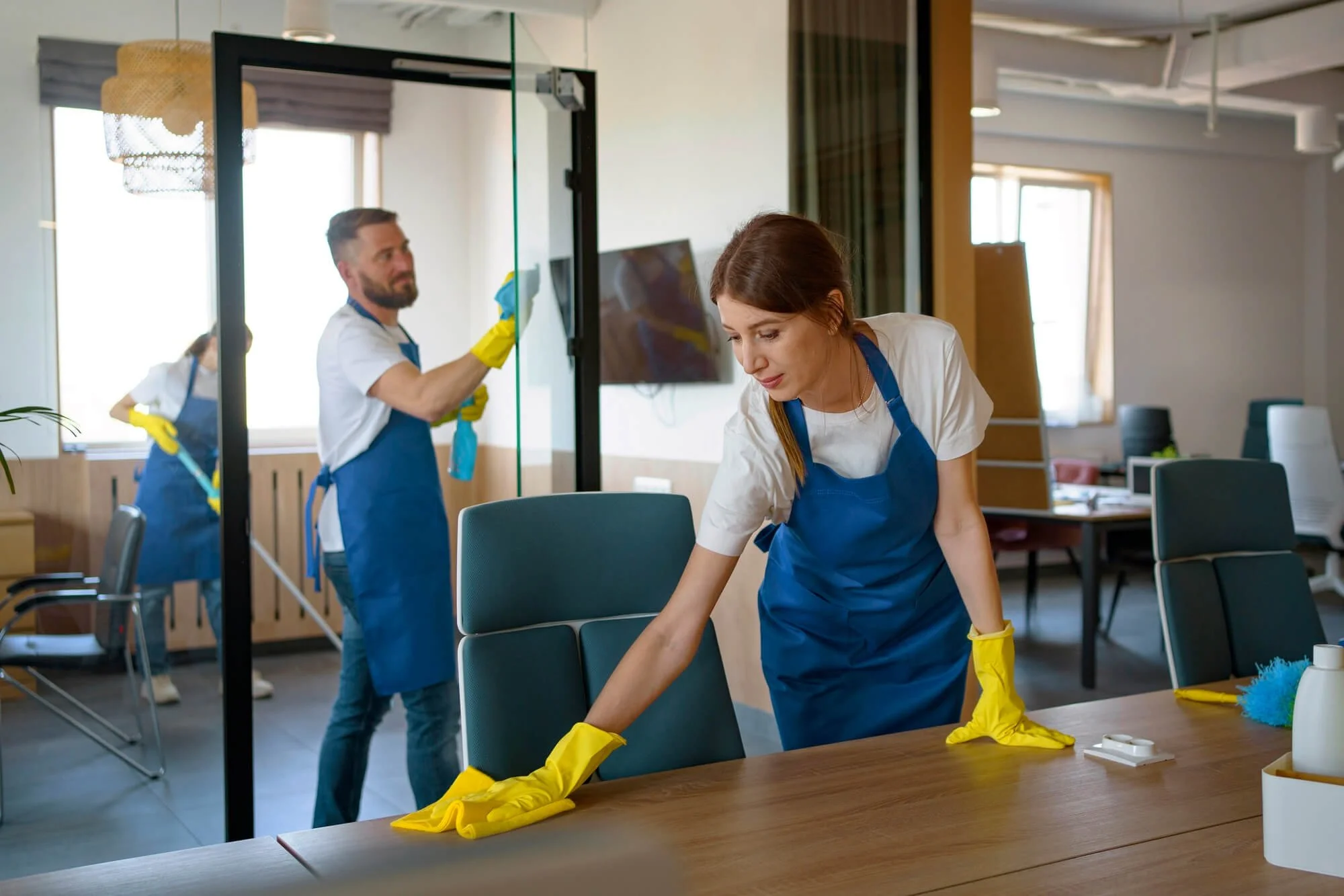 Three people cleaning an office. The woman in the front is wiping a table, the man in the background is cleaning a glass partition, and another woman in the back is mopping the floor. They are all wearing blue aprons and yellow gloves.