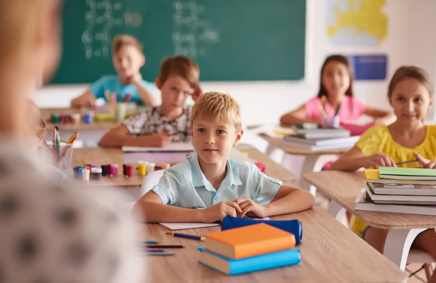 A classroom full of young students sitting at desks with school supplies, paying attention to a teacher at the front. There is a green chalkboard in the background.