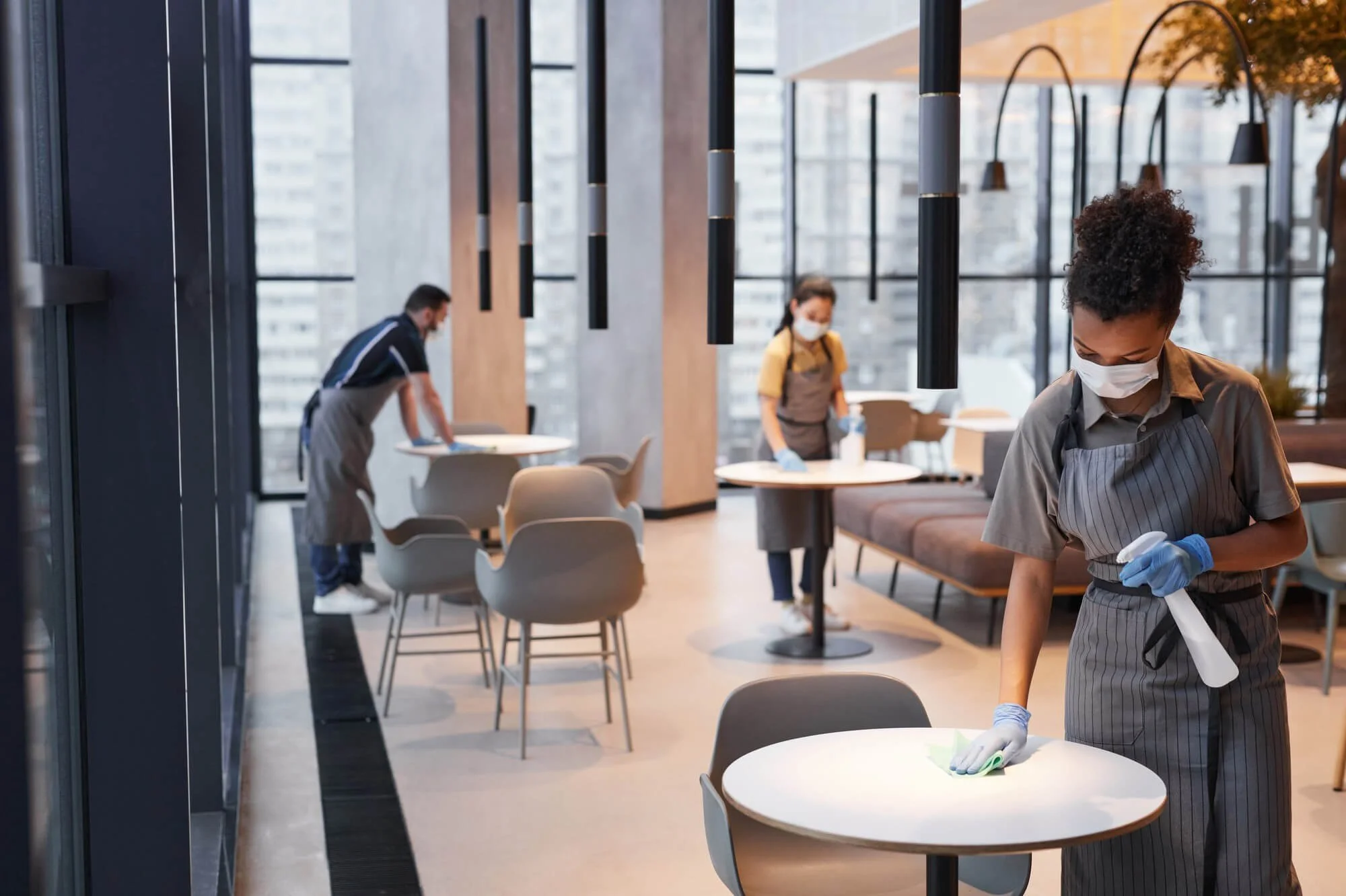People wearing face masks and gloves cleaning tables in a modern restaurant or cafe with large windows and high ceilings.