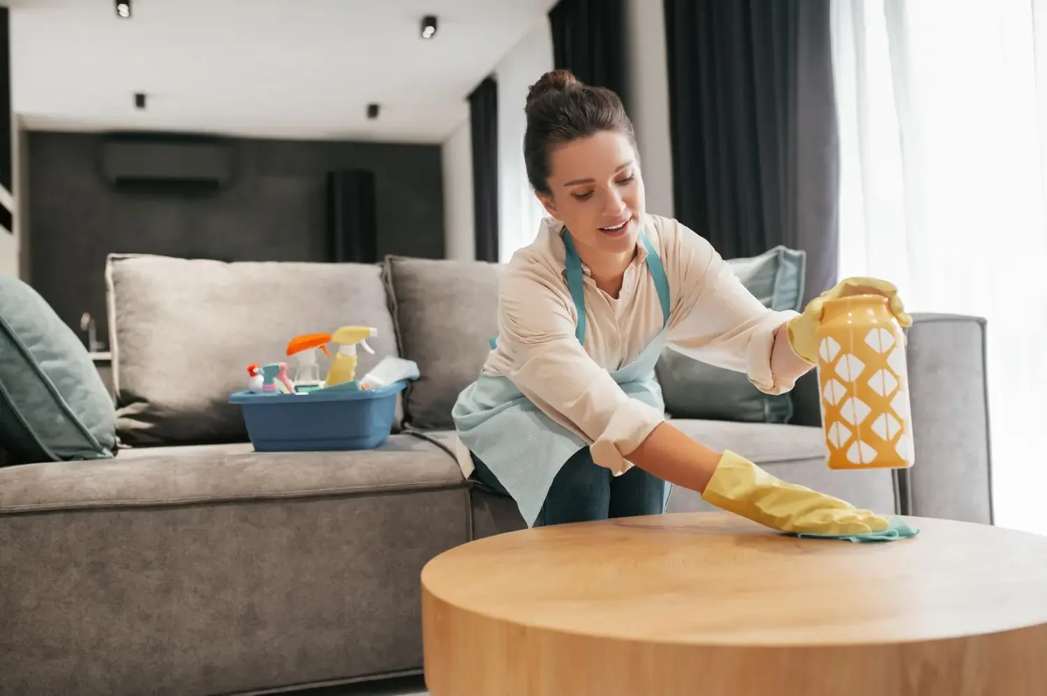 Woman cleaning a wooden coffee table in a living room, wearing yellow rubber gloves and an apron, with cleaning supplies in a blue container on the gray sofa behind her.
