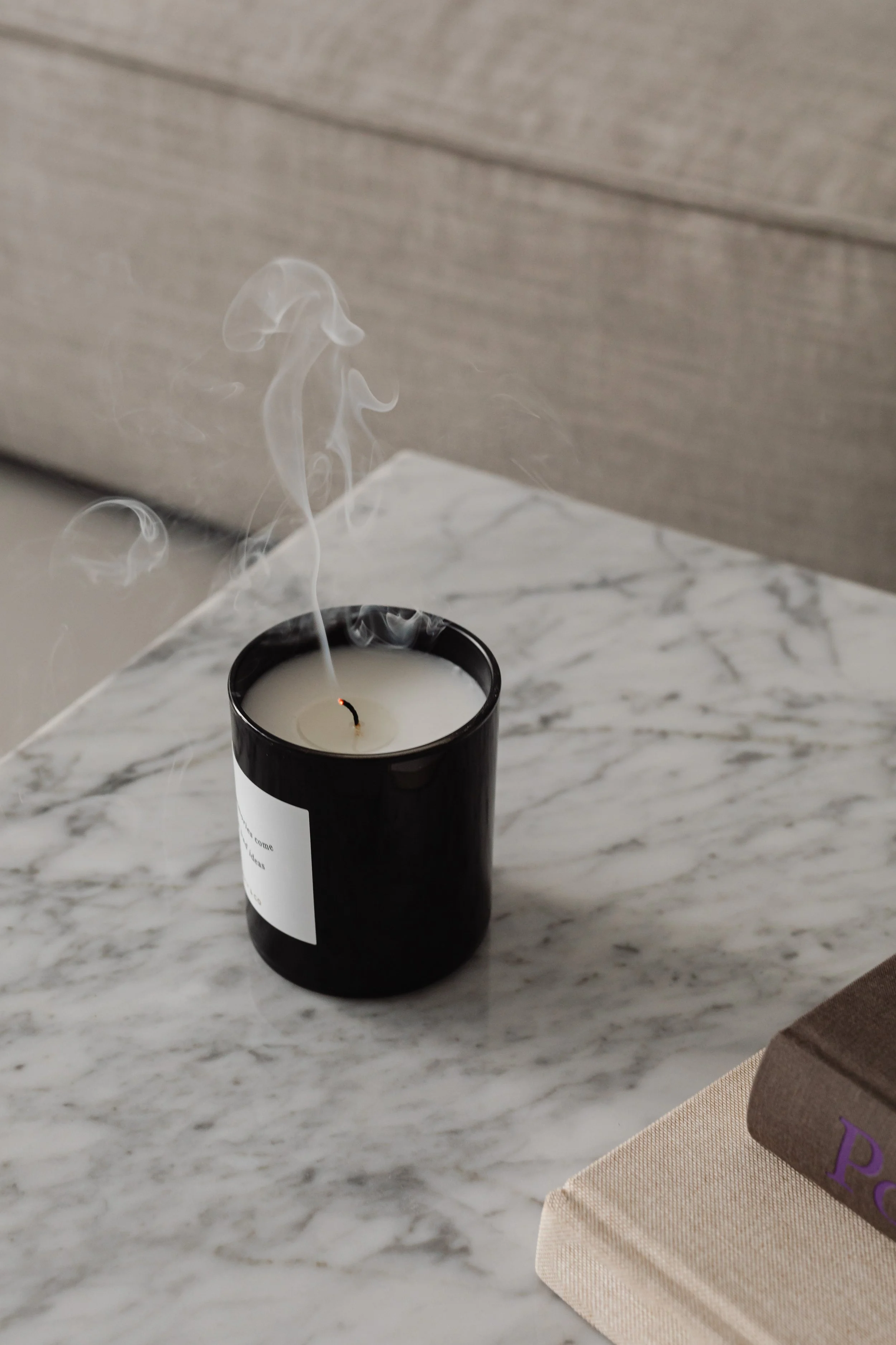 A black scented candle with smoke rising from the wick, placed on a marble surface beside a beige and a brown book.
