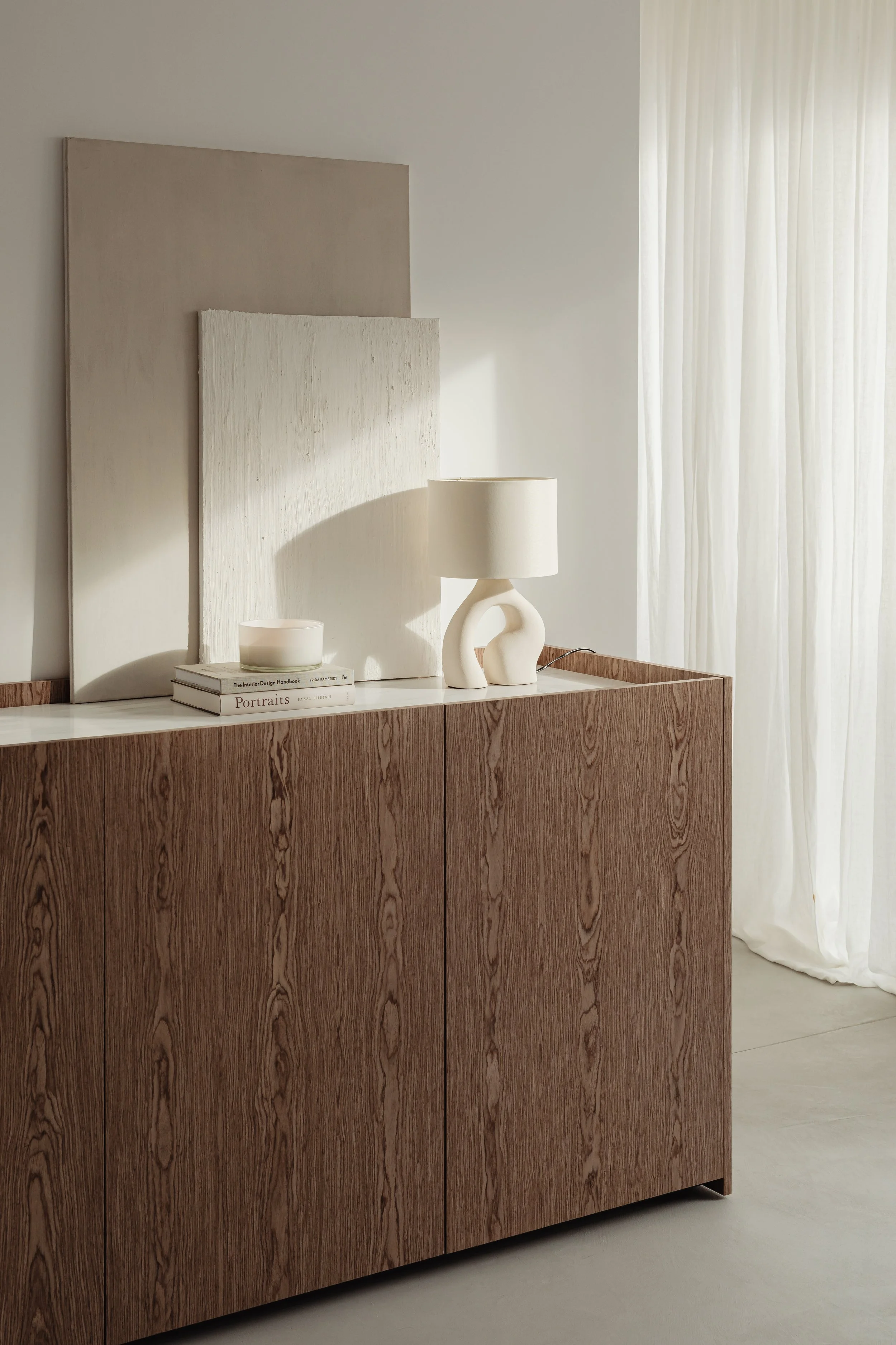 A wooden sideboard with a white top, decorated with a white ceramic bowl, books, and a modern white table lamp with a sculptural base. Behind the sideboard, a large abstract artwork leans against the wall. White curtains cover a window, allowing soft natural light into the room.