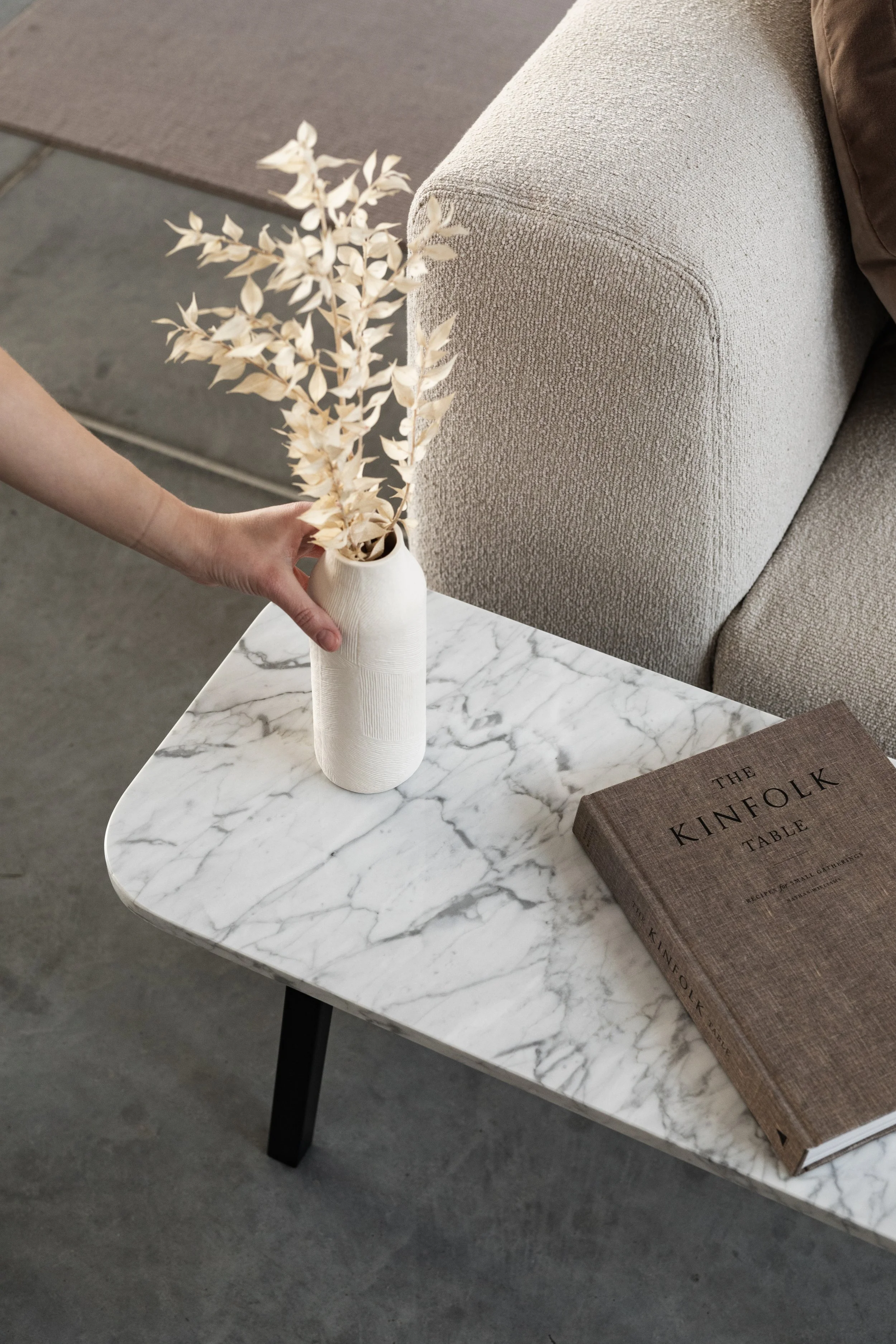 Person's hand placing a white vase with dried beige leaves on a white marble side table next to a hardcover book titled 'The Kinfolk Table' in a modern living room.