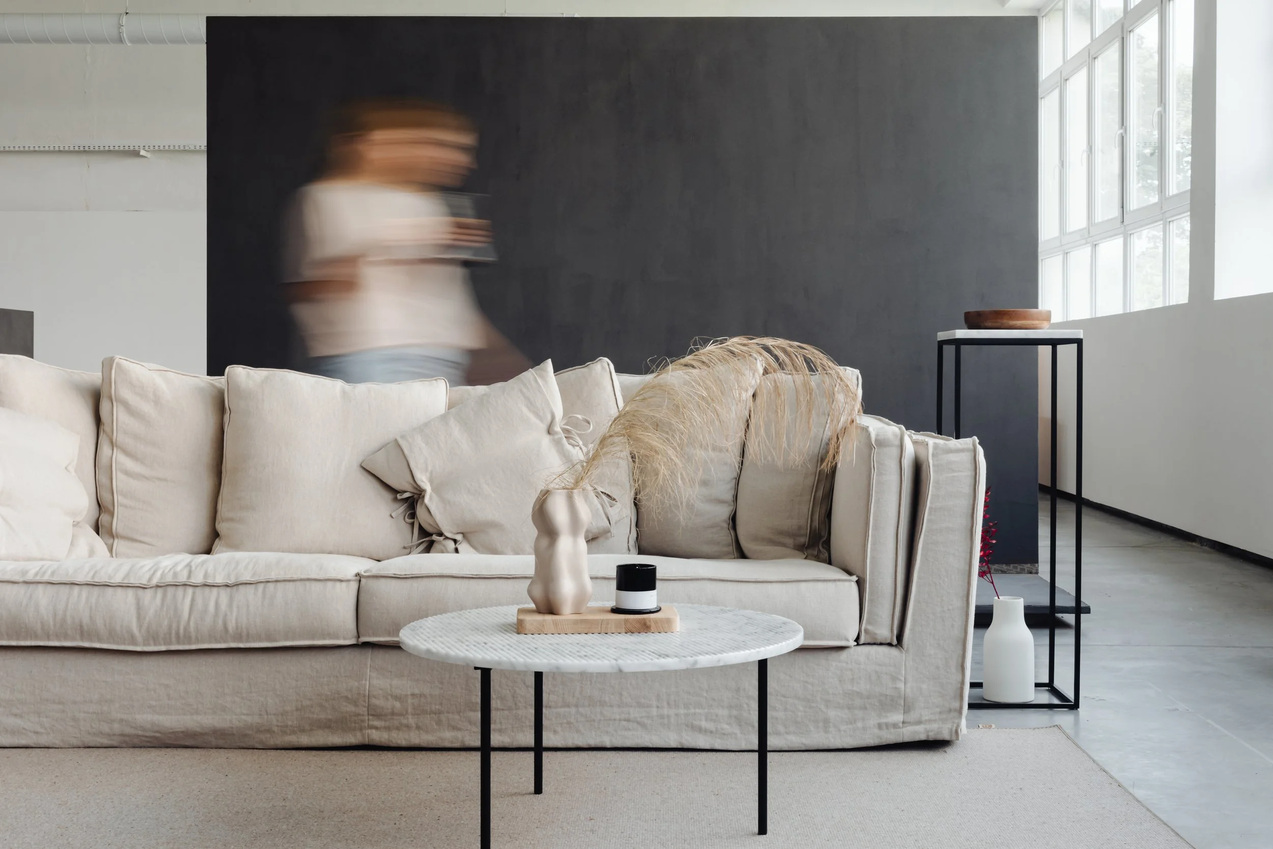 Living room with beige sofa, white cushions, and a round white coffee table, hosting decorative vases, with a black wall and large windows letting in natural light.