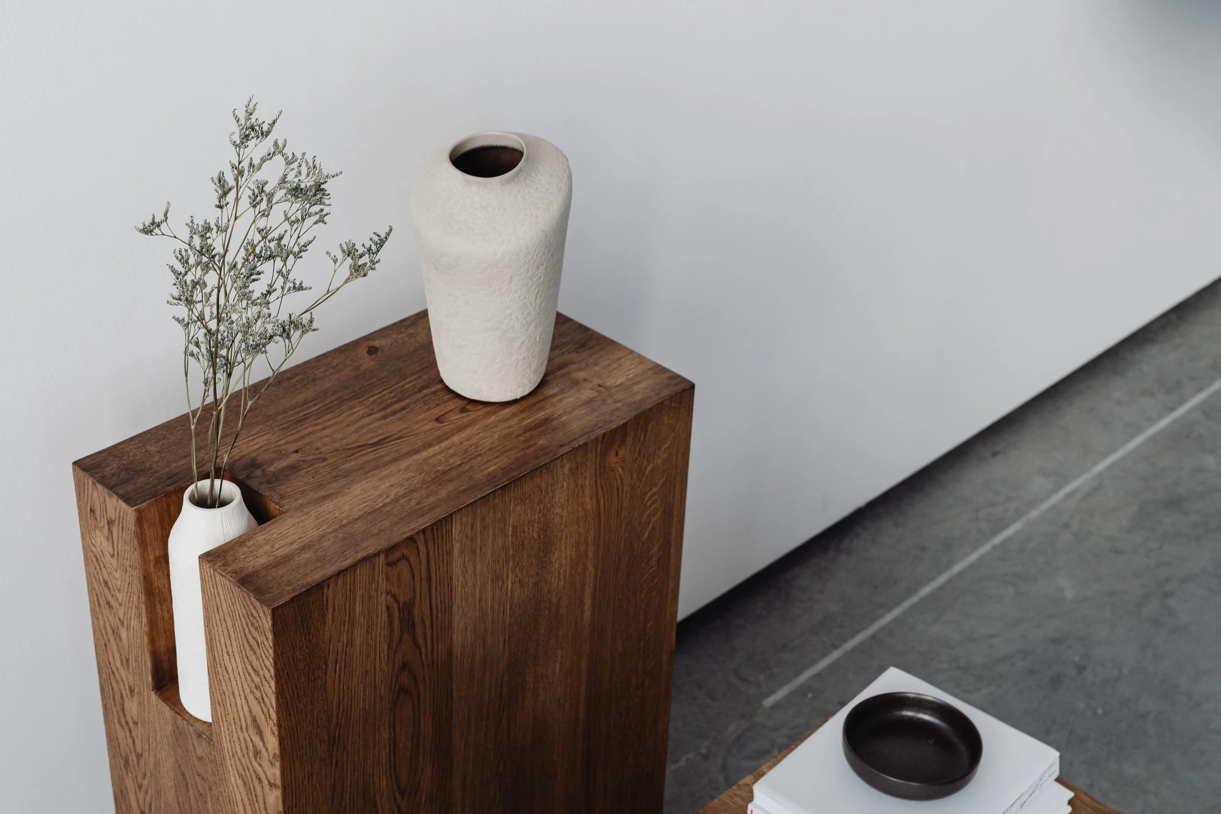 Decorative interior with a tall white textured vase and a smaller white vase with dried flowers on a wooden pedestal, and a black dish on a stack of books on a wooden surface, against a plain white wall.