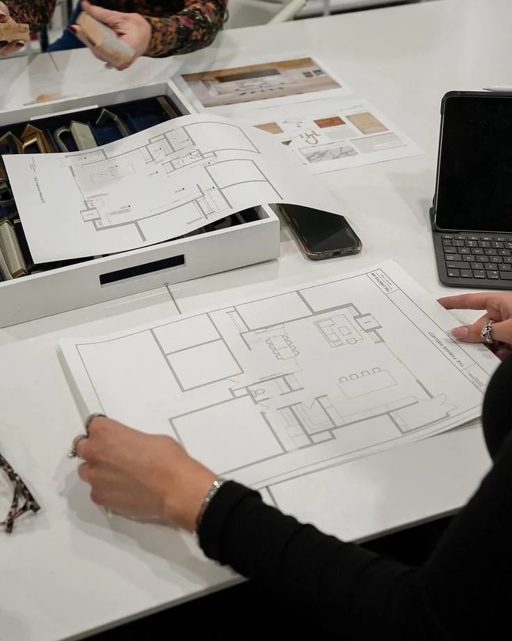 Design plans and blueprints of a house on a desk, with color samples, a smartphone, and a tablet nearby, during a planning or design meeting.