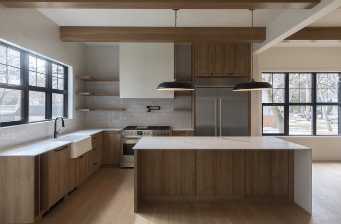 Modern kitchen with large windows, wooden cabinets, a white island, and stainless steel appliances.