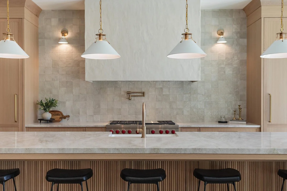 Modern kitchen with a marble island, black bar stools, a stove with red knobs, pendant lights, and minimal decor including a plant and candlesticks.