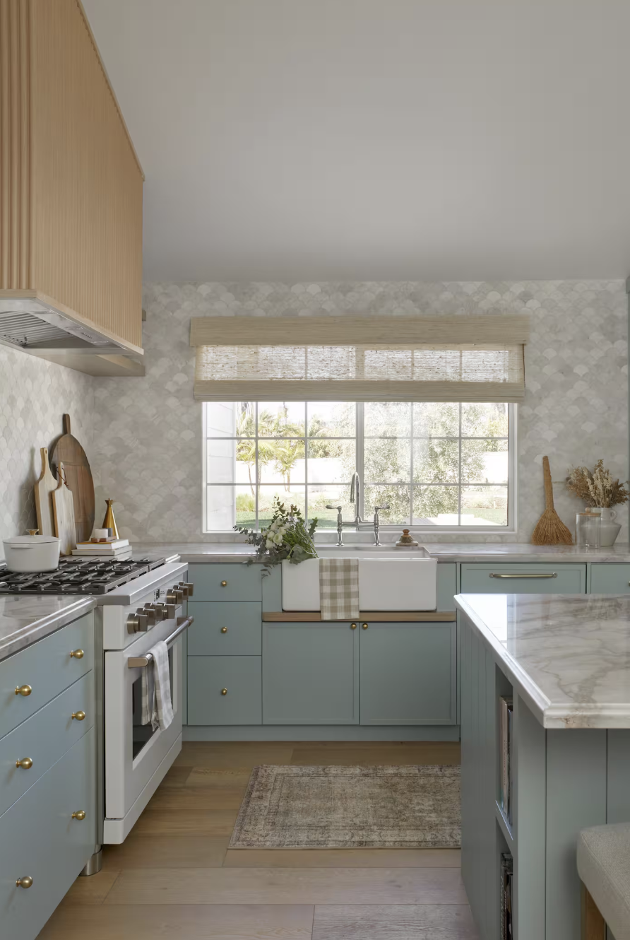 Bright kitchen with blue cabinets, white marble countertops, a farmhouse sink, and a large window showing outdoor greenery.