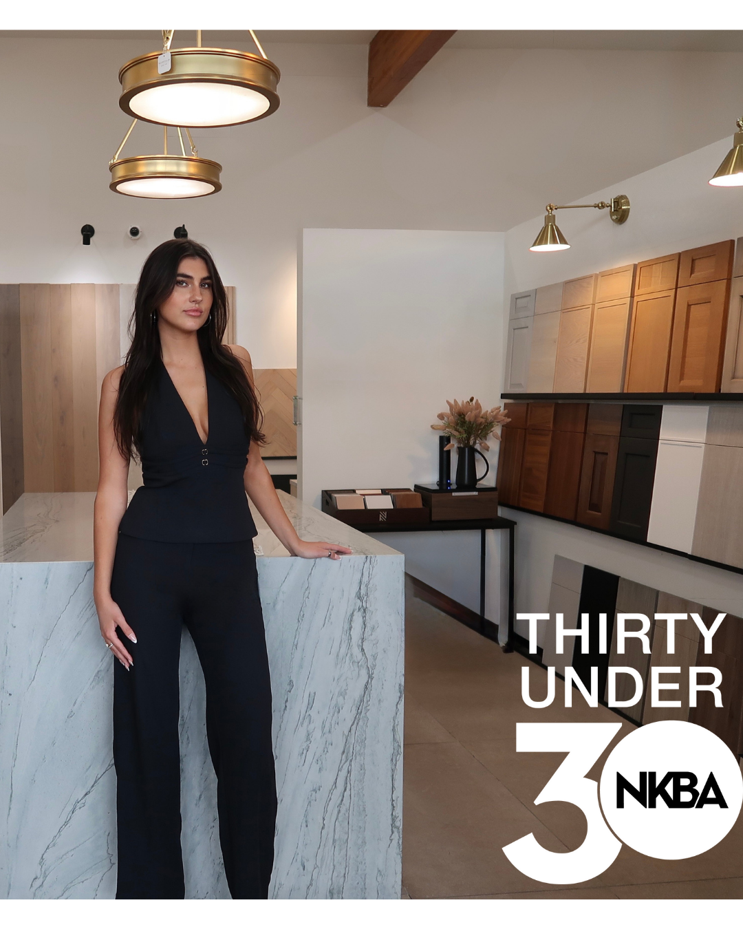 A woman in a black outfit standing next to a marble counter in a modern kitchen with wooden cabinets. There are pendant lights and a decorative vase with flowers on the counter. The image includes the text 'THIRTY UNDER 30 NKBA'.