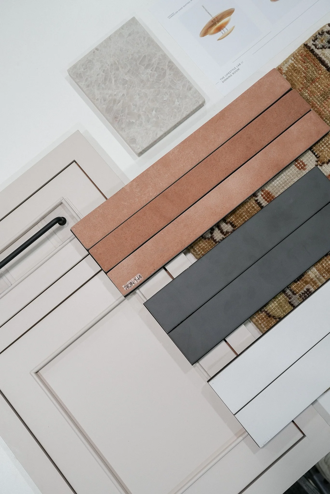 Display of various tile samples in colors such as pinkish, dark gray, and white, along with a textured beige tile and a white cabinet door. Some samples are labeled, and the setup appears to be in a showroom or store.