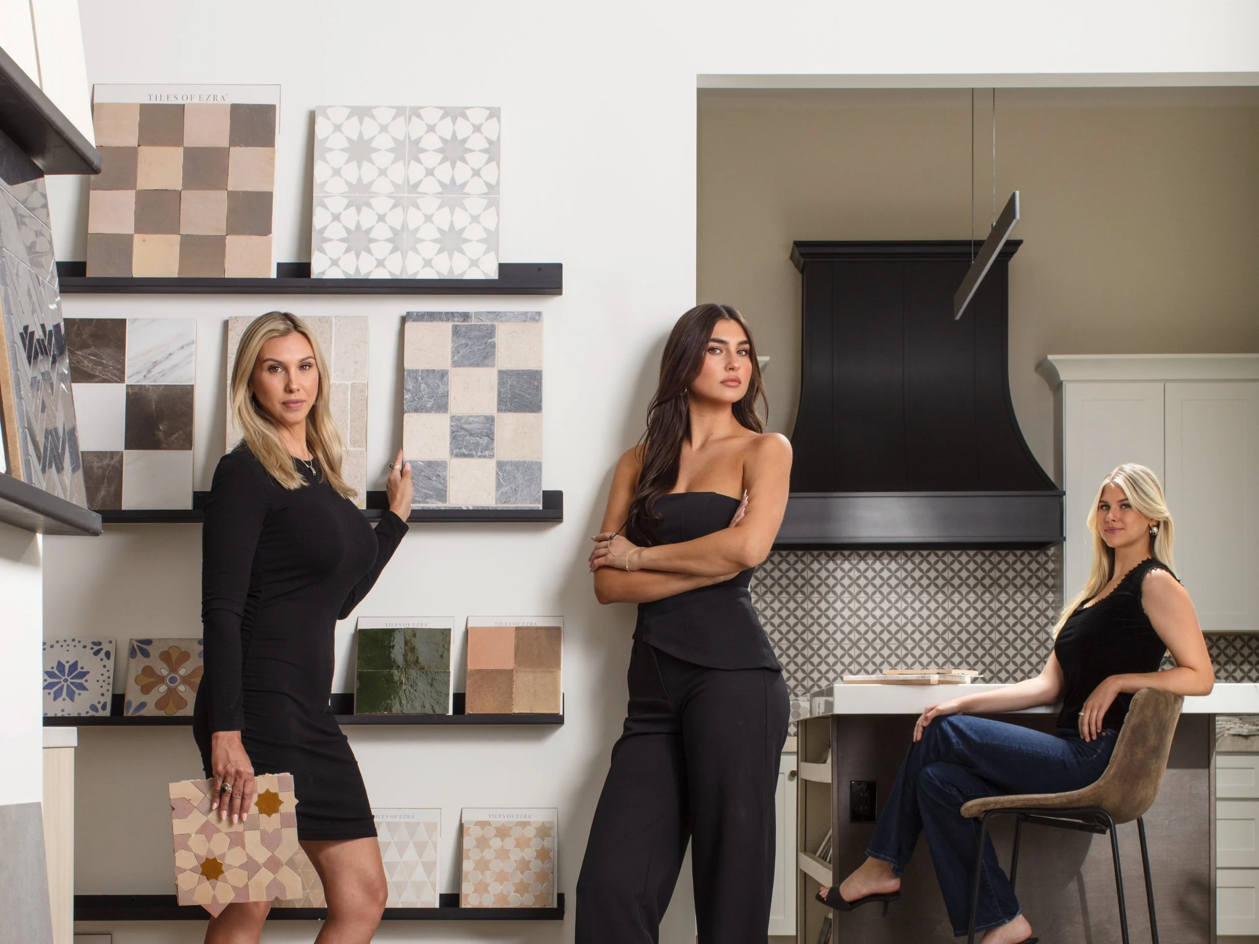 Three women in a tile showroom, with various tile samples on display. Two women are standing, and one is seated on a chair.