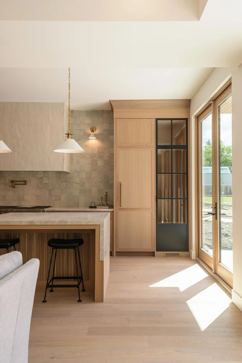 Modern kitchen with light wood cabinetry, a marble island, and large glass doors allowing sunlight to fill the space.