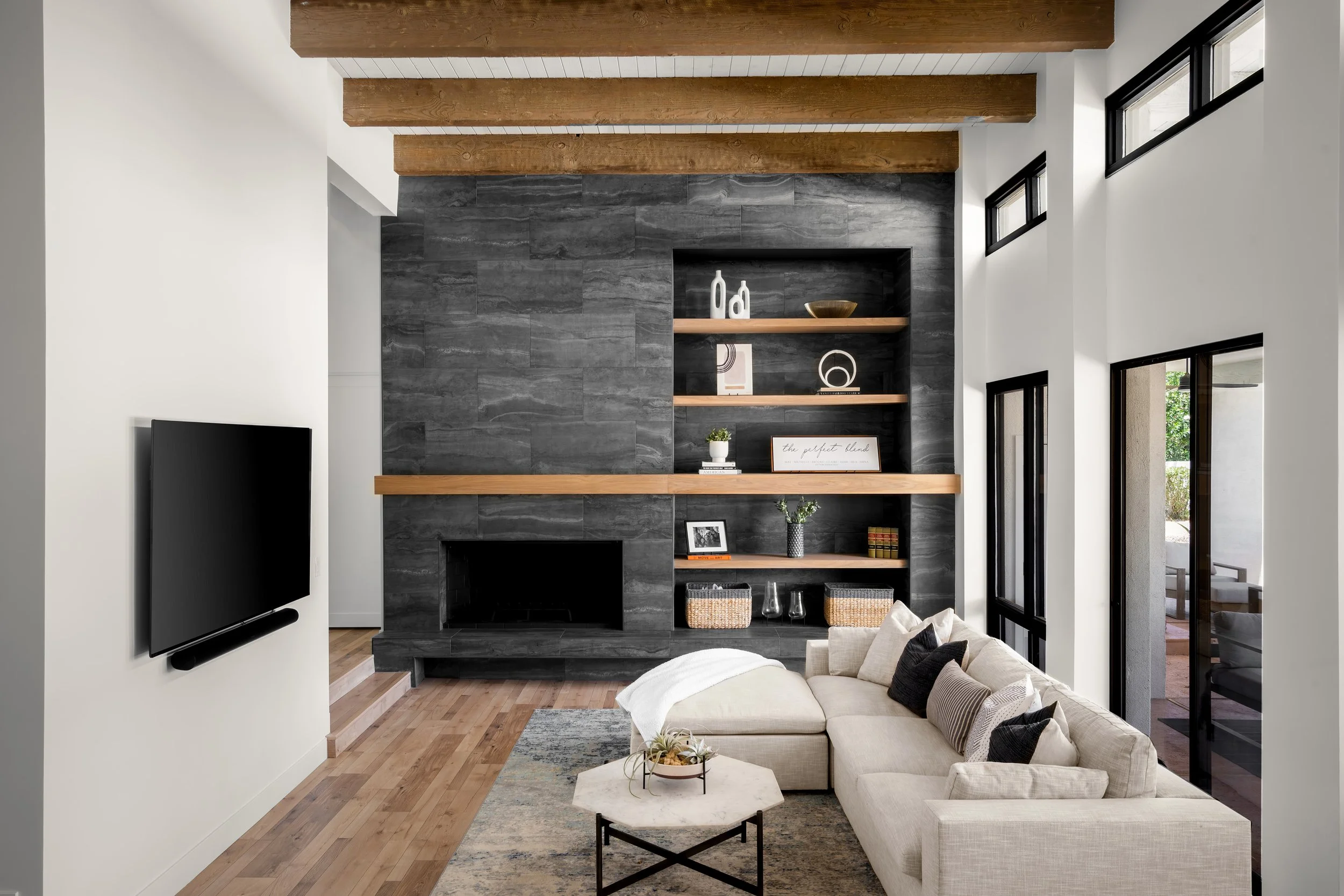 Modern living room with a built-in dark stone fireplace, wooden shelves with decorative items, cream sectional sofa, and large windows.