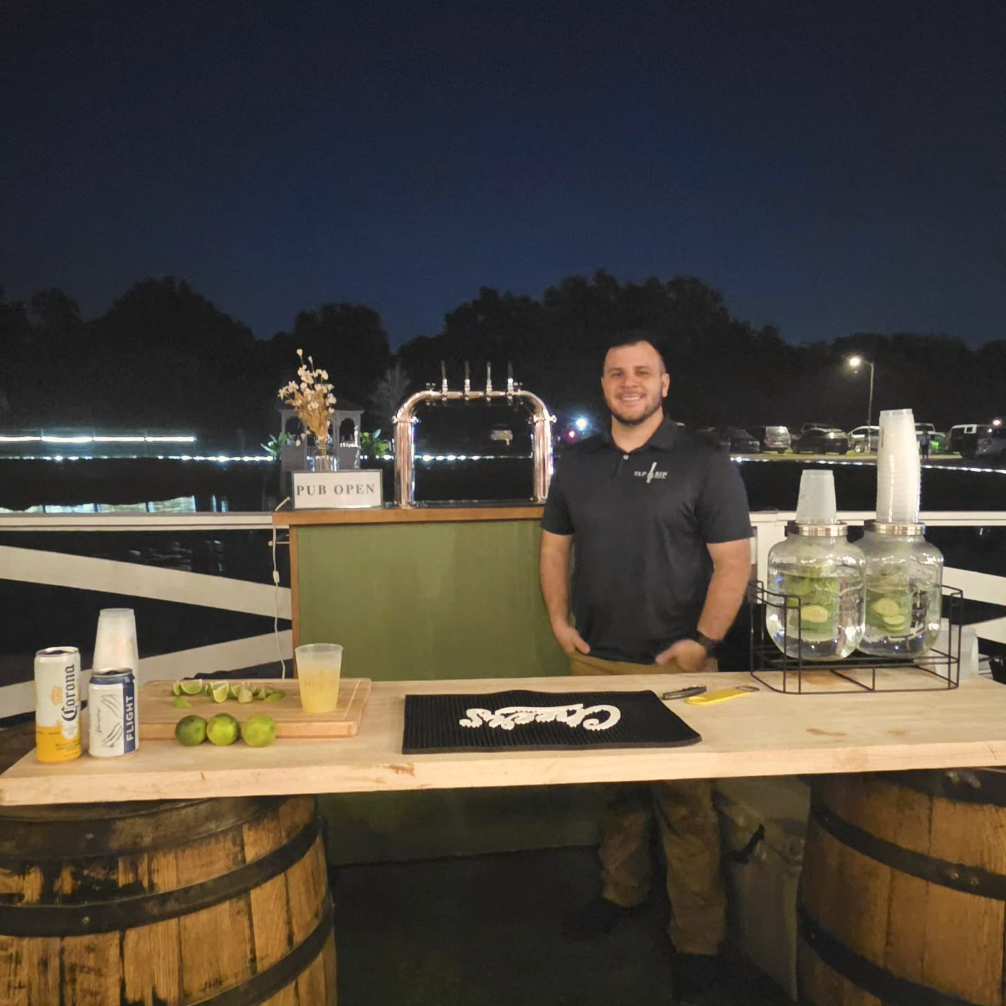 A man standing behind a wooden bar counter decorated with limes and mixers, smiling at the camera at an outdoor venue during nighttime, with a sign that reads 'PUB OPEN'.