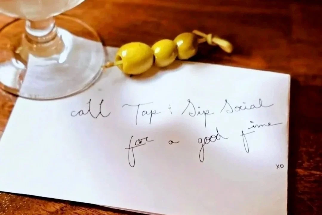 A handwritten note on a piece of paper that says, 'call tap & sip social for a good time.' There is a wine glass and some yellow berries or decorative items on the paper, sitting on a wooden surface. a martini bar and smoked drinks and garnishes 