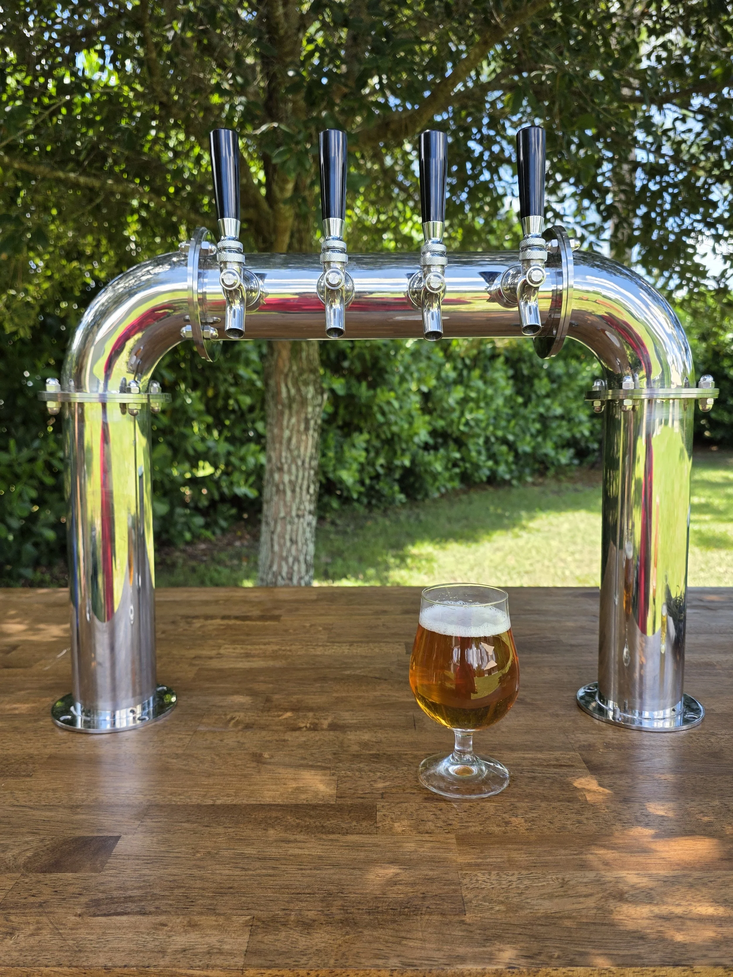 A wooden outdoor bar table with a short glass of beer and a four-tap beer dispenser, set against a background of green bushes and a tree.