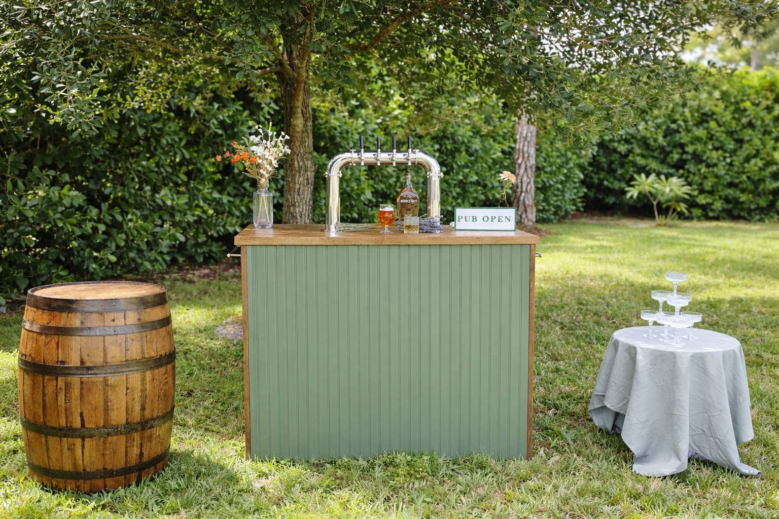 Outdoor pub bar setup with a wooden barrel, a green bar with a beer tap, a sign that reads 'PUB OPEN,' a vase with flowers, a bottle of alcohol, a glass of beer, and a table with a champagne tower, set in a grassy area with trees and bushes in the background.