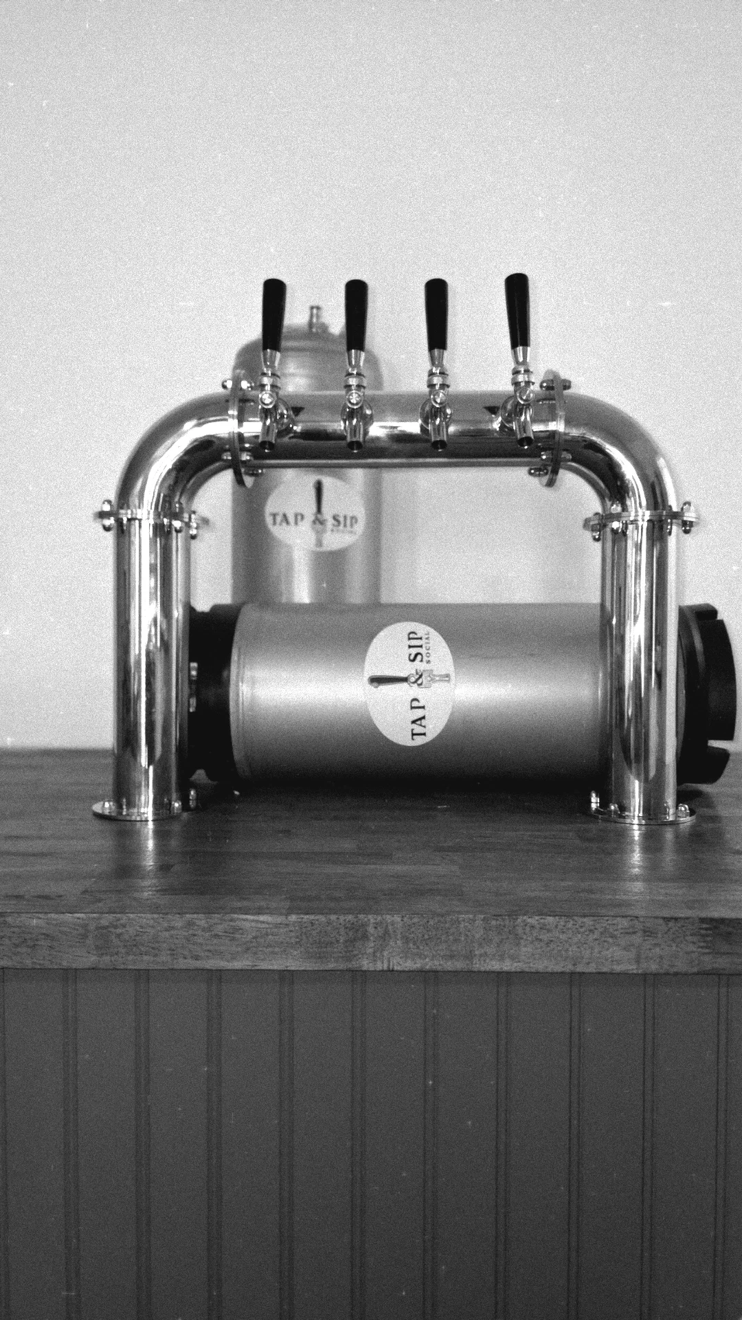 A stainless steel beer tap dispenser with four taps, labeled 'Tap & Sip,' situated on a wooden counter against a plain wall. A custom mobile draft cart so its a mobile bar and with cocktail kegs and draft beer