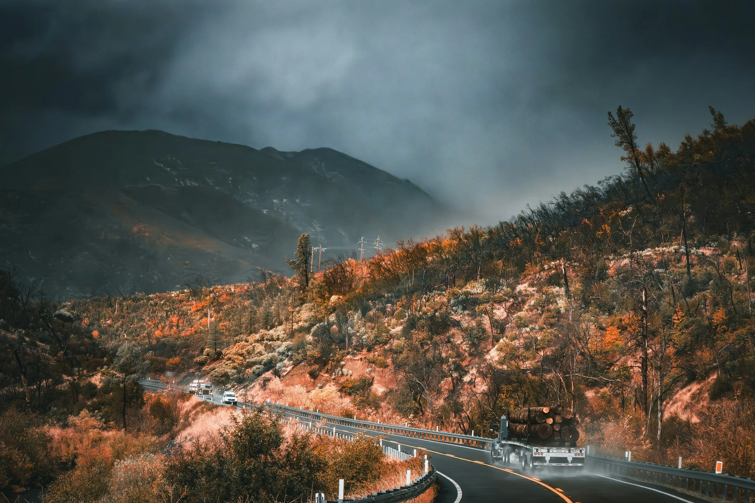 A winding mountain road surrounded by autumn foliage, with a truck carrying logs and a line of cars traveling in the rain under dark, cloudy skies.