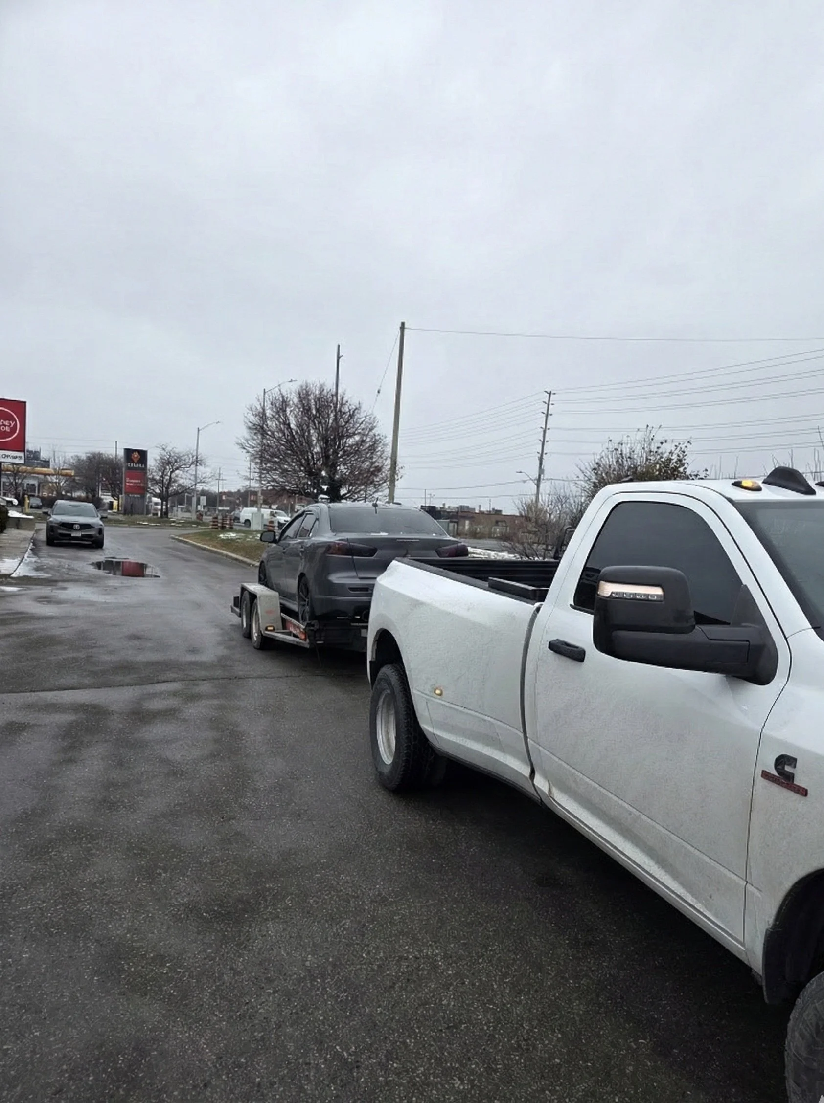 White pickup truck towing a black car on a trailer in a parking lot on a cloudy day.