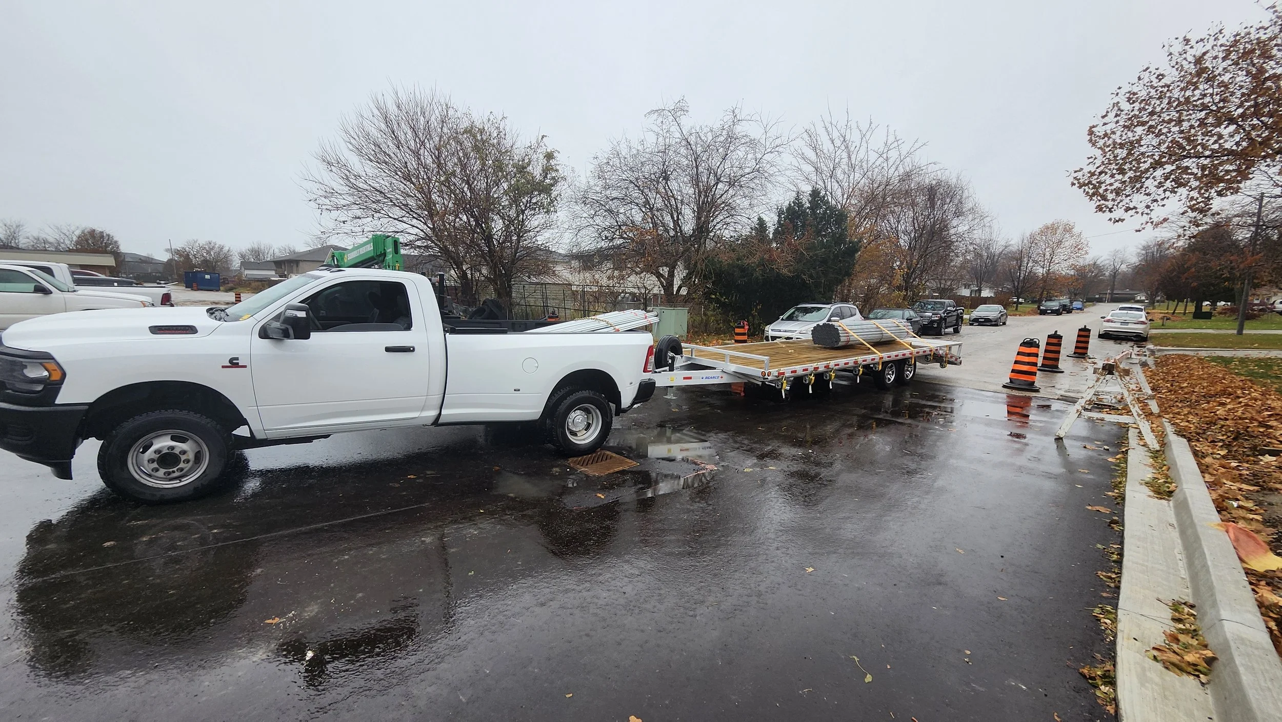 A white pickup truck with a flatbed trailer attached, carrying rolled-up wooden planks, parked on a wet street on a rainy day, with orange traffic barrels placed along the roadside.
