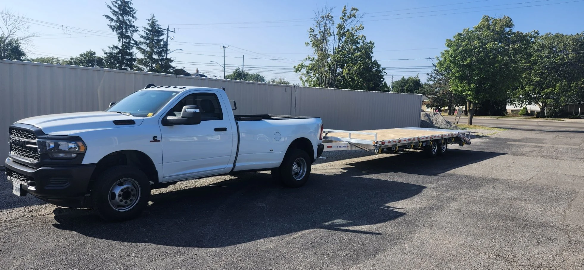 White pickup truck with a flatbed trailer parked on a paved lot, with trees and a metal fence in the background under a clear blue sky.