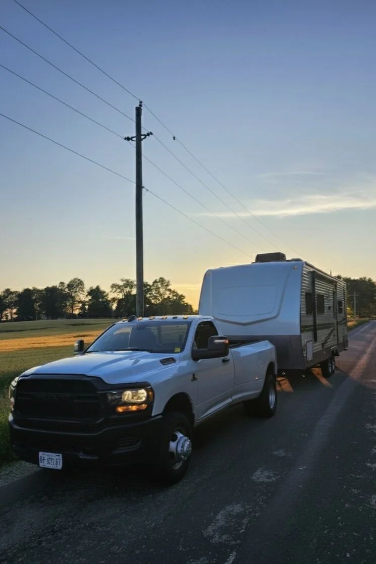 A white pickup truck towing a travel trailer parked on the side of a rural road at sunset.