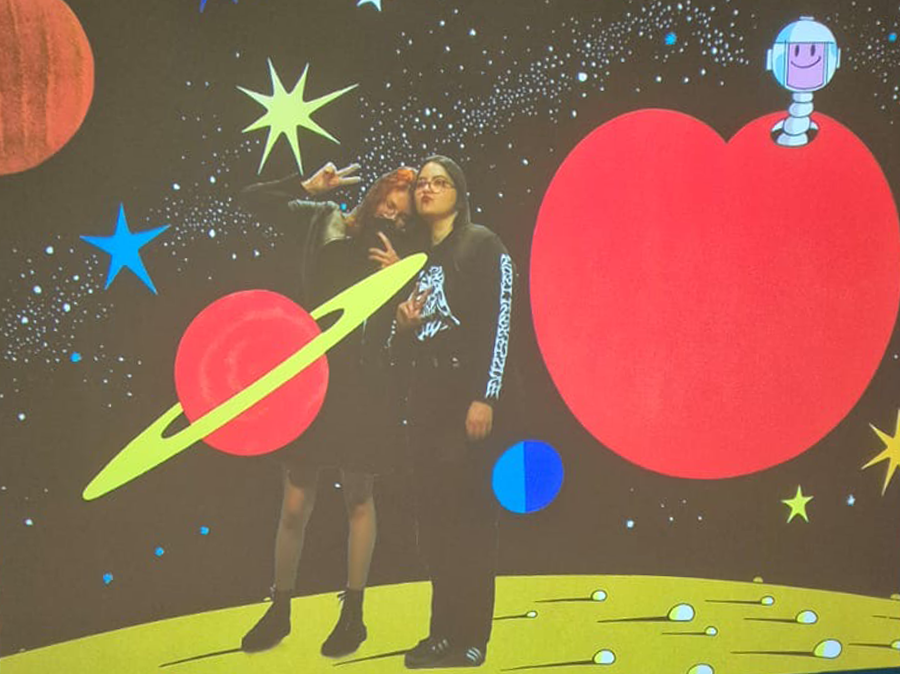 Duas pessoas posando em um fundo com tema espacial, incluindo planetas, estrelas e uma nave robótica em formato de maçã, com cores vibrantes e desenhos de espaço.