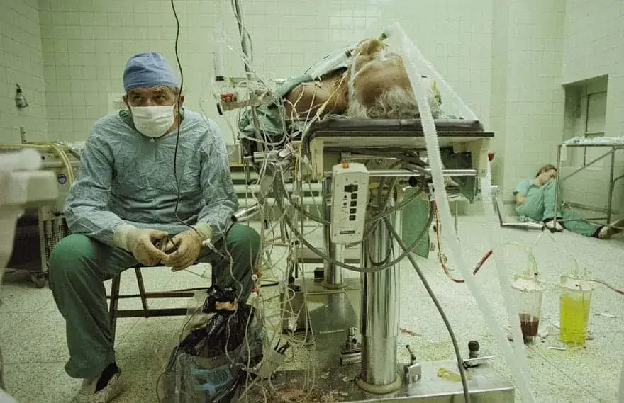 A medical professional in scrubs and a mask sitting next to a patient connected to a ventilator in an intensive care unit.