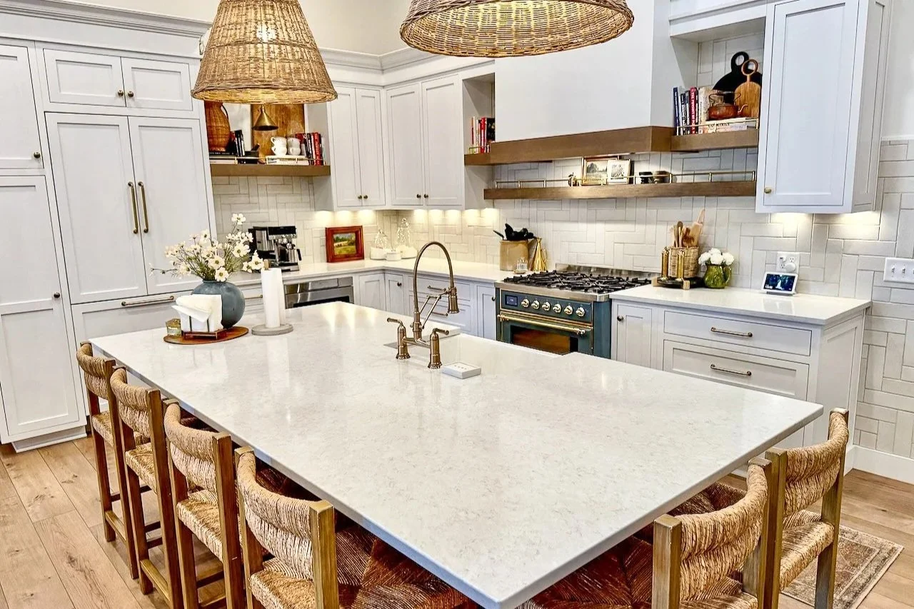 Modern kitchen with white cabinetry, a large central island with a white stone countertop, and woven pendant lights. Features include a blue oven, open wooden shelves with books and decor, and a vase with flowers on the island.